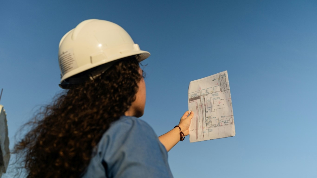 A person in a hard hat holding and looking at a construction plan against a clear blue sky.